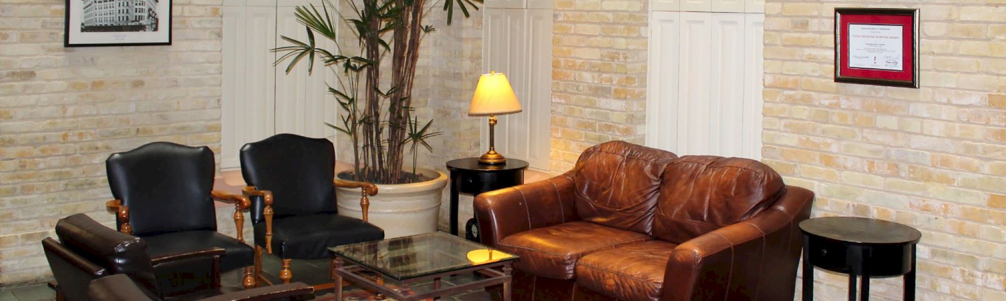 A cozy lobby or waiting area with a brown leather sofa, two black chairs, glass coffee table, lamp, plant, and framed art on a light brick wall.
