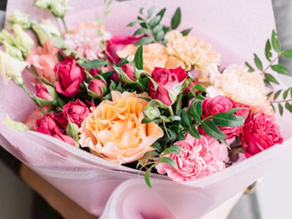 A bouquet of colorful roses and carnations in pink, peach, and coral, wrapped in light pink paper, with greenery accents.