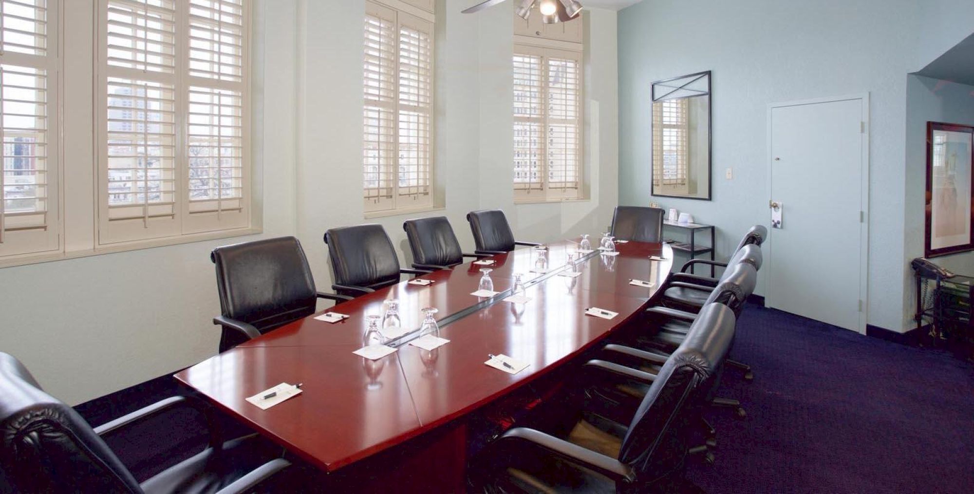 A long conference table with black leather chairs in a bright room, windows with blinds, and place cards or papers at each seat.