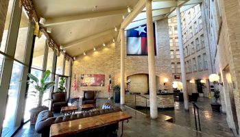 A stylish hotel lobby with tall windows, stone floors, industrial beams, a long wooden table, cozy seating, plants, and a Texas flag hanging high.