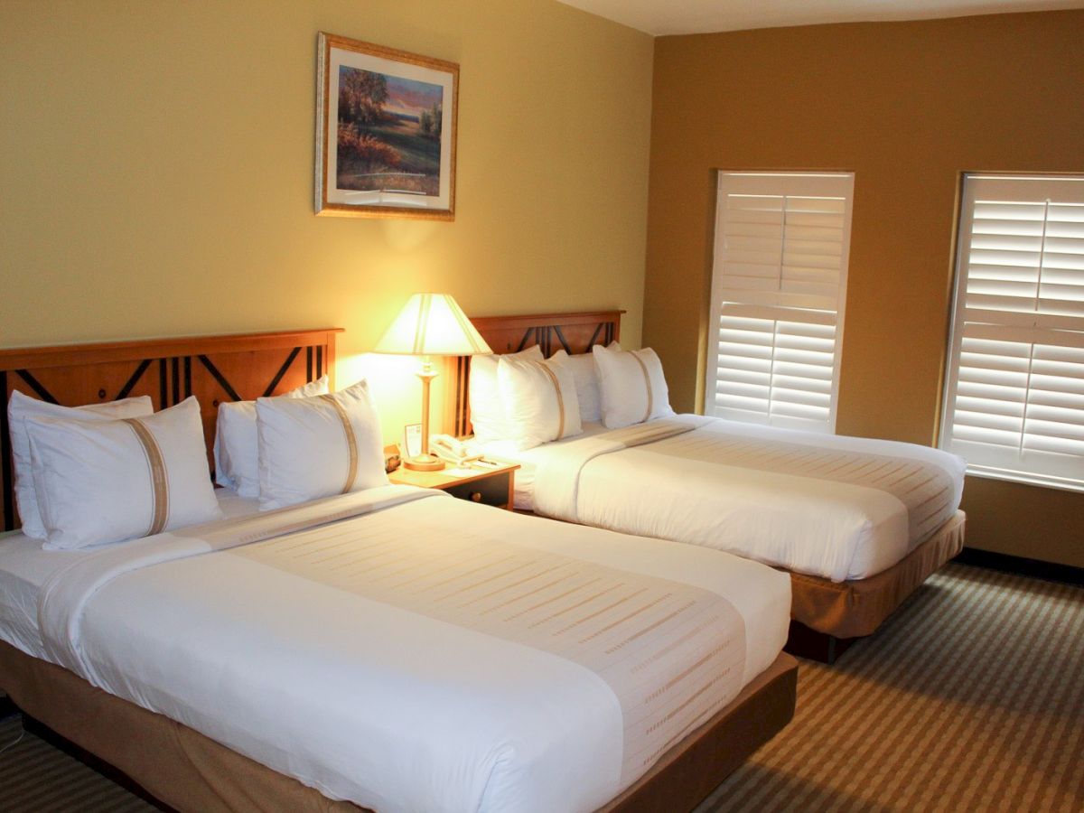 A hotel room with two double beds, nightstand with lamp, framed art, and beige walls with white blinds—cozy and tidy.