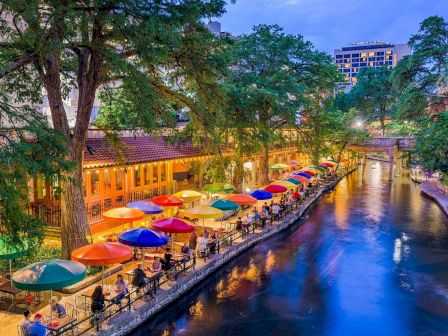 Colorful canalside dining with umbrellas and warm lights reflecting on the water; people stroll along the riverside terrace at dusk.