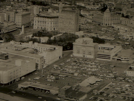 A black-and-white cityscape with low-rise buildings, a grid of streets, and a large parking lot in the foreground, showing an urban downtown area.